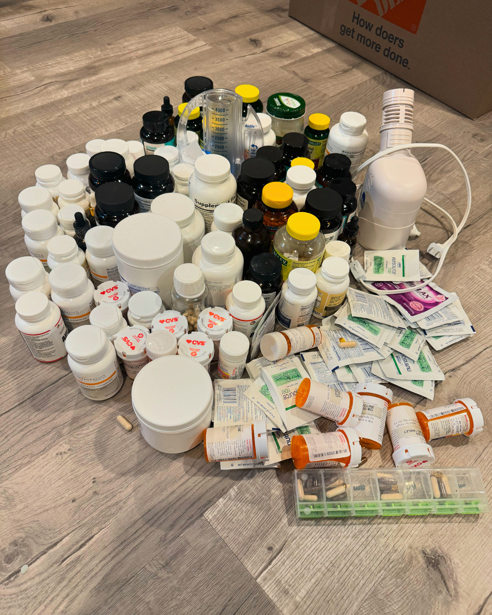 Bottles, packets, and a pill organizer of medicines and supplements spread on a wood floor